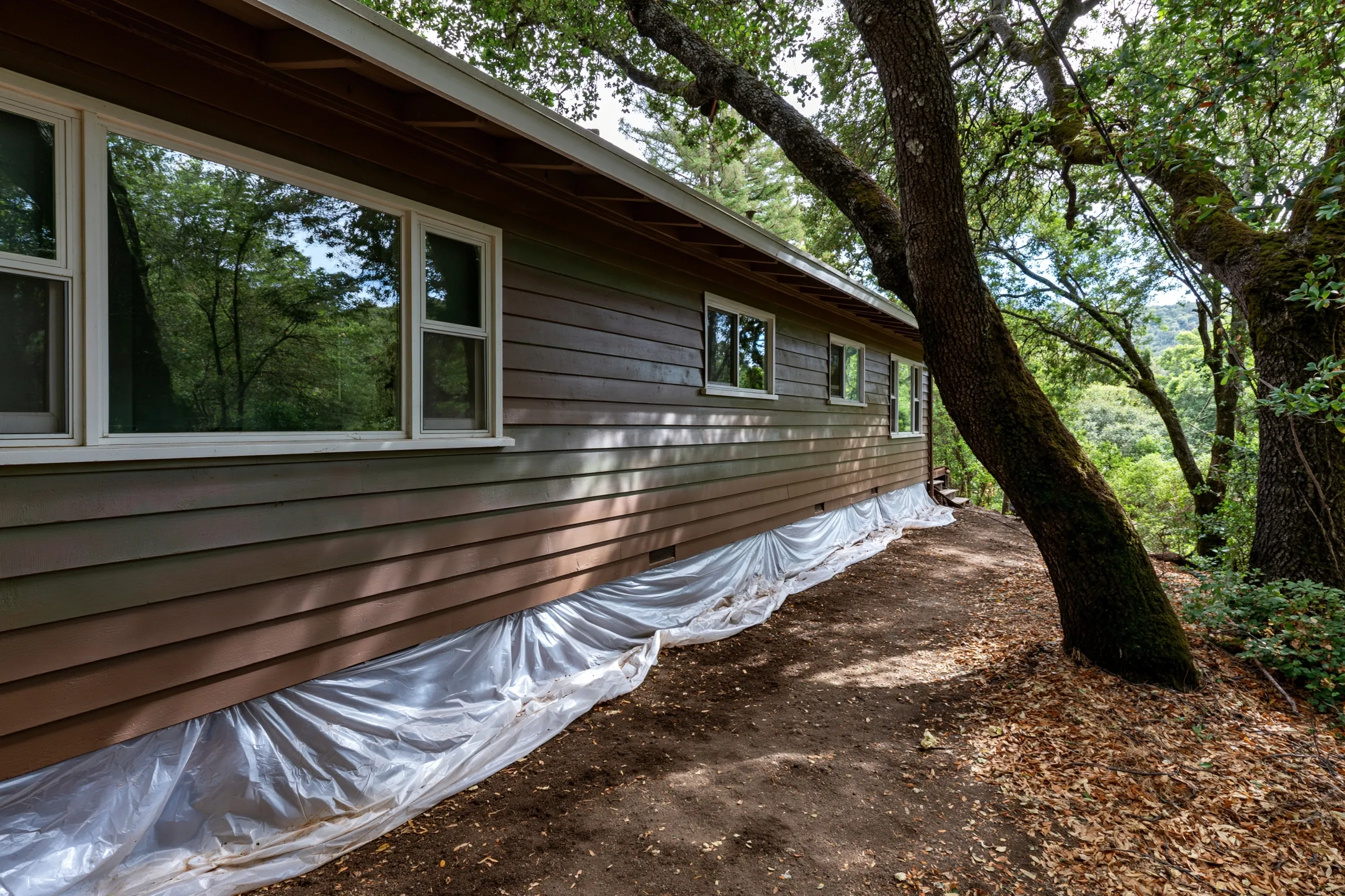 Long ranch house with horizontal wood siding under oak trees, plastic sheeting spread on the ground along the wall